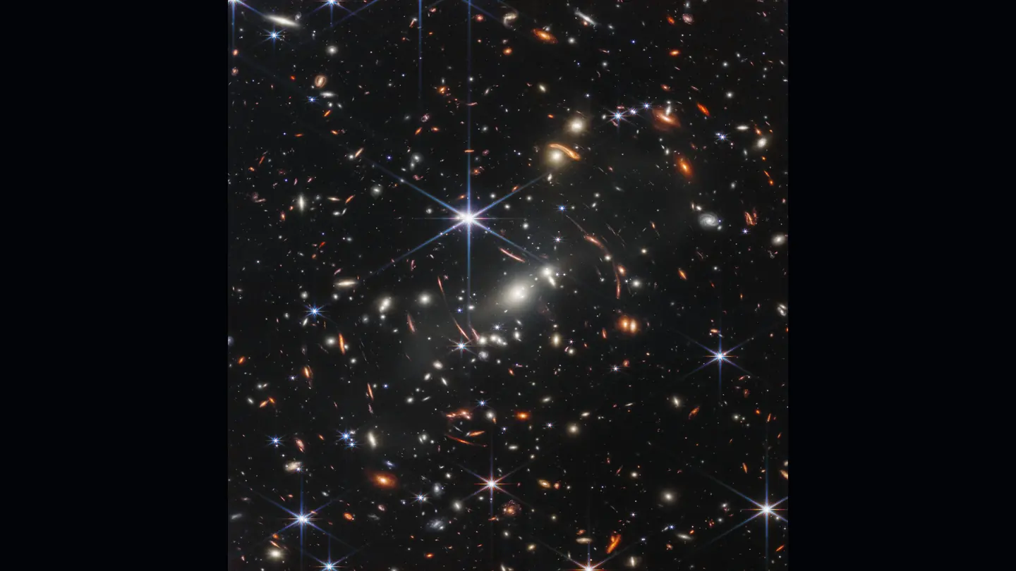 Webb deep field filled with thousands of galaxies, bright diffraction-spiked stars, and thin curved gravitationally lensed arcs.